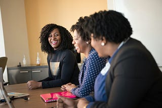 Three women sit together at a conference table engaged in conversation, smiling and laughing during a meeting. Laptops and notebooks are visible on the table, and the atmosphere conveys collaboration, leadership, and teamwork in a professional setting.