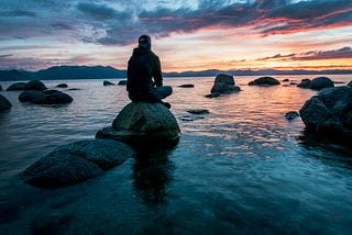 A picture of a man sitting on a rock and enjoying the serenity of the sunrise on a lake.