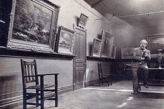 Black and white photo of an art gallery with classic paintings in elaborate frames, a simple wooden chair and a man sitting thoughtfully in the corner.