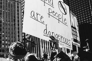 A group of protestors is gathered on a city street holding a sign saying “Stupid People are Dangerous”