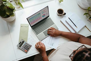 A busy solopreneur works on a laptop at a white desk cluttered with a calculator, documents, a green envelope, a cup of coffee, and a spiral notebook with a pencil on top. Small green plants add a touch of nature to the organized chaos. Image credit: Microsoft 365 (Unsplash) — “Solopreneur sitting at a table with a laptop” under the following license: Unsplash Free to Use.