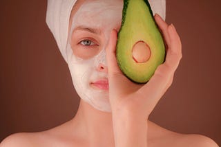 A woman holds an avocado infront of the left side of her face, while the right side is covered in white beauty cream.
