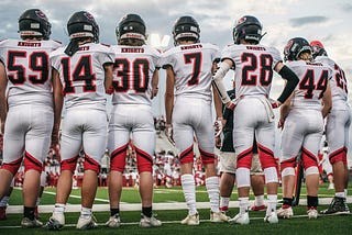 Several American football players stand with their backs to the camera, listening to instructions from their coach