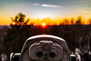 In-focus coin operated binoculars overlooking a blurry backdrop of the sunset and large trees.
