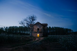 A haunting two-story farmhouse in a desolate wood. The front door is open, light on in an otherwise dark farmhouse.
