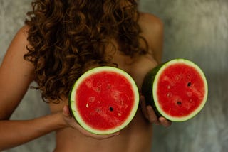 A woman holds two halves of a watermelon against her, like breasts. Cut open, as we are once we have breast cancer.