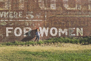 A little sassy girl standing in front of a brick wall with the words, where, 5, and for women are spray painted on it.