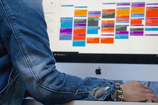 E-calendar view on Apple desktop. They are color-coded and time blocked. A person’s arm and hand are in the foreground — it appears that they’re scheduling an event on this calendar.