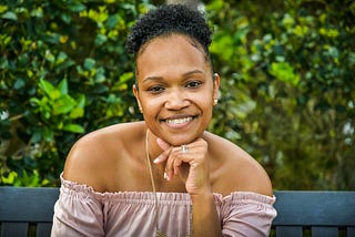 Woman smiling and sitting on a bench behind a bush and other green vegetation. They are wearing a pink top.