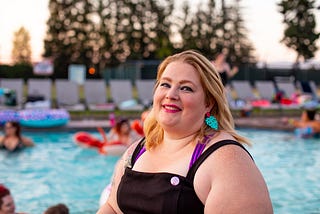 In the foreground is the top half of a fat woman in her thirties who is standing in front of a large in-ground swimming pool. She is wearing a black retro style swimming costume and dangly, bold, aqua colored earrings. Her head is slightly tilted back and she has a gentle smile. In the background people are swimming and sitting on colorful inflatables. The sun is setting and there is a row of beige pool loungers.