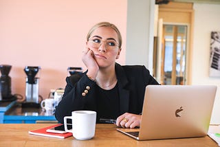 woman with glasses and pantsuit at table with coffee and laptop, head leaning on hand, thinking look.