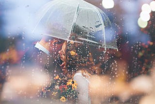 A man and a woman standing close to each other under umbrella because it is raining. The woman is whispering something in man’s ear.