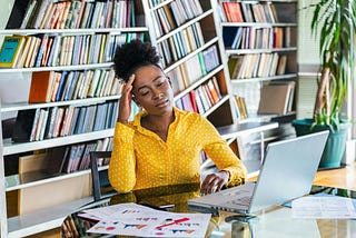 A Black woman at her computer looking stressed with her eyes closed and fingertips touching her forehead.