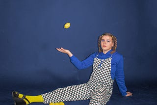 Lady in chequered dungarees over a blue sweater and yellow socks, sitting on the floor, juggling one lemon with others on the floor around her.