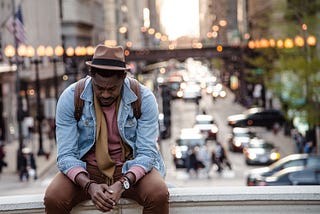 A man sitting with his head bowed down, traffic and city lights behind him.