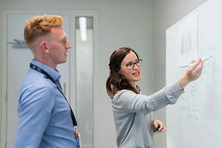Young woman pointing at a white board and a young man watching the whiteboard standing next to her