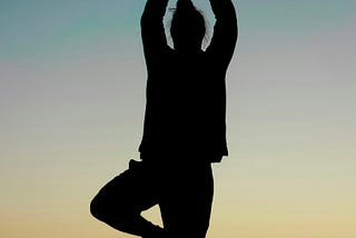 Silhouette of someone performing the ‘tree’ yoga pose, with the sunset behind her.