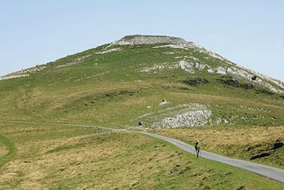 A flat-topped green, treeless, and rocky hill dominates the scene. A bit of pale blue sky. A narrow grey path bisects the scene, and a distant, lone walker heads toward the viewer.