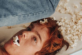 A picture of a boy lying on the bed with popcorn in his mouth, others in his hair and on the surface around him. The image depicts getting lost in the popcorn