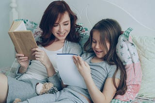 A mother and daughter relax together on a bed, reading, with smiles on their faces.