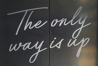 Steel elevator doors prepare to open so you can rise to greater elevation. Written in upbeat cursive across the metal: “The only way is up.”