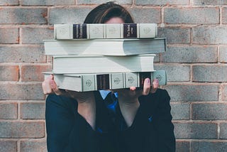 A woman holding books up to obscure her face. She stands in front of a brick wall.