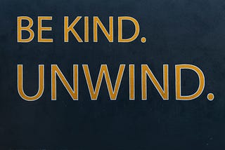 The words “Be kind. Unwind.” written in gold on a dark background.