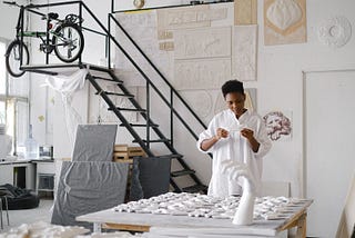 Woman in a white art studio, wearing a white dress and hold a piece of plaster shaped like a bird. The table infront is full of plaster mouldings and the walls are covered in flat sculptures.