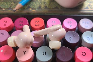 brightly colored keyboard close-up with pale pink earbuds on top and and a green notebook in the background. a blue pencil is sitting on the green notebook.