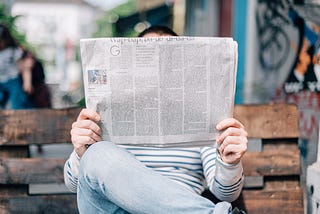 A photo of someone sitting on a wooden park bench, with their legs crossed, holding up a broadsheet newspaper. The newspaper covers their face; only the top of their head, with short black hair, is visible