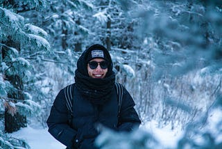 Smiling person in dark jacket, hood, and glasses, in front of snow-covered trees