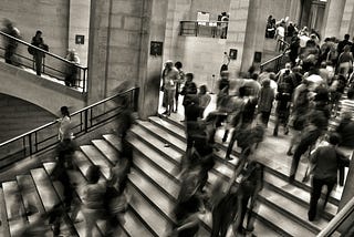 This image depicts a bustling interior scene, possibly within a museum or public building, characterized by a sense of motion and activity. The central focus is a grand staircase populated by numerous individuals depicted in various states of motion blur, which suggests the dynamic flow of people moving up and down. Some individuals are captured in stillness, offering contrast to the blurred motion around them, which gives the scene a temporal dimension. The architecture features classic stone w