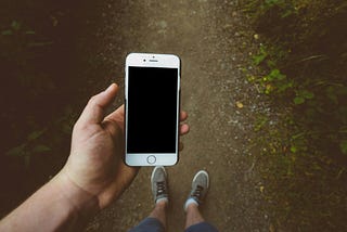 A person standing on a trail in the woods (view only of legs with feet clad in socks and sneakers ) looking at an empty screen on a smartphone.