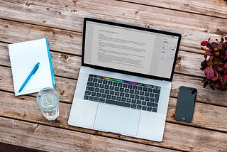 Macbook on wooden table showing resume on screen, plus notebook, pen, phone, plant and glass of water next to it