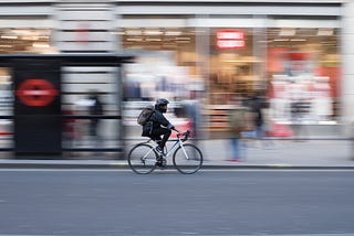 A photo of a cyclist going down a city street, taken from the side, with the cyclists heading to the right, and the shops in the background blurred by motion