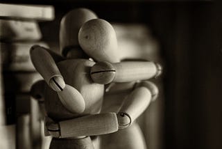 Sepia toned image of two wooden artist’s mannequins wrapped in an embrace