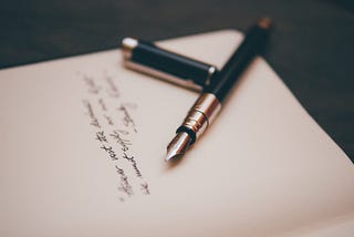 An open diary with cursive text written in it. Lying on the page is an open fountain pen with a golden nib.