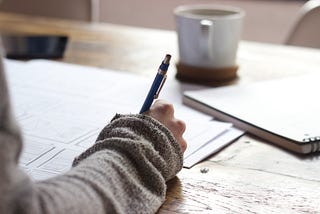 Person’s arm using pen to write on paper. Coffee cup and notebook on desk top.
