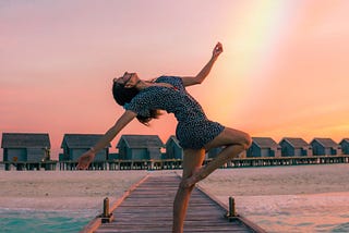 Woman on a dock of a beach posing in dance with a rainbow in the background behind her at sunset.