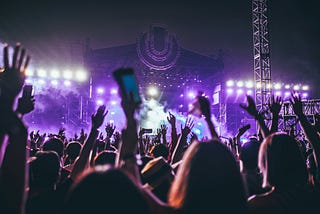 Crowd at a concert faces stages, arms in the air, purple stage background