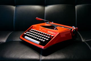 A red old-fashioned typewriter on a black leather couch.