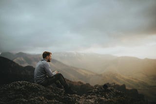 A man sitting on top of a mountain next to a forest.