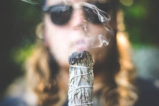 Image of burning sage in front of a woman’s face wearing sunglasses.