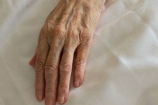 The wrinkled, blue veined hand of an old woman lies atop white sheets.