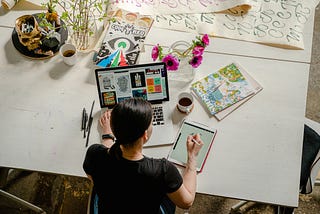 Photo of a woman writing on her tablet while also using a laptop. The desk contains various art materials, implying that she’s creative and artistic. The lighting is soft, and a sense of calmness pervades the scene, suggesting that the woman is focused and in a state of flow.