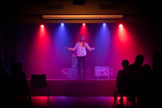 A white man with a beard wearing a white collared shirt, red scarf, and dark pants stands on a stage backlit by alternating blue and red lights in an otherwise dark room. He has his arms slightly outstretched. The dark silhouettes of a few people in the audience can be seen.