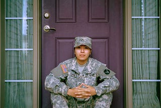 A woman soldier gazes pensively into the foreground from a doorway, where she is sitting and relaxing.