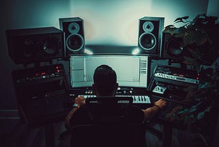 Someone sitting at a computer with speakers above the screen. They have a musical keyboard in front of them and recording equipment on either side