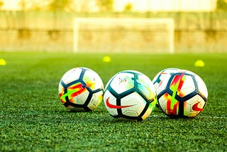Three soccer balls sit on a turf soccer field with small yellow cones and a goal blurred in the background.