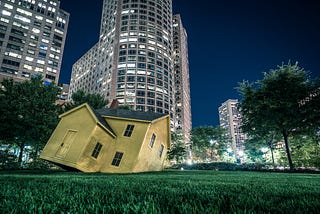 Obra de arte que representa uma casa amarela instalada num parque urbano em meio a prédios. A casa está desnivelada ao solo.